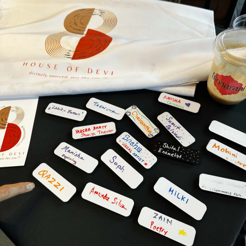 Name tags with various names and professions are laid out on a black surface in the foreground, with a white t-shirt featuring a logo and a drink in a plastic cup visible in the background.