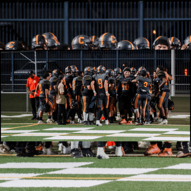 A football team huddles on the field in front of a fence lined with helmets, with the legs of players visible in the foreground.