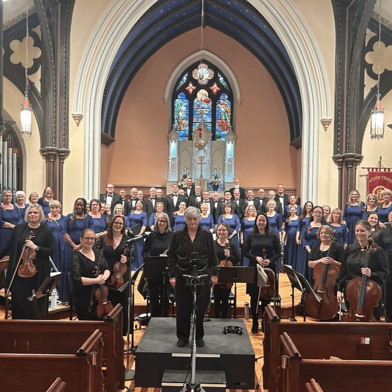 A choir and orchestra stand in a church with stained glass windows and ornate architecture in the background.