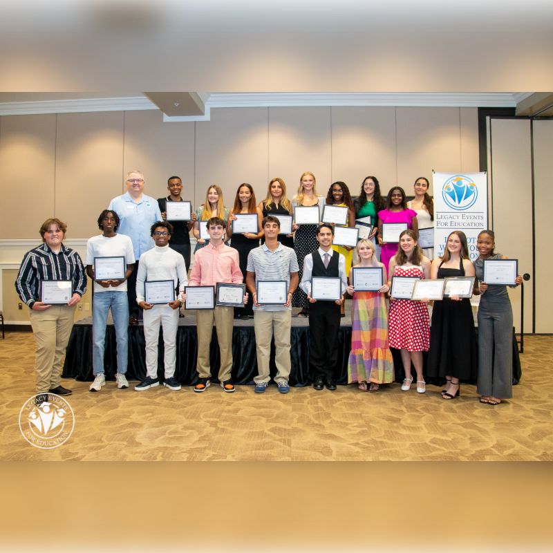A group of students and an adult stand on a stage holding certificates, with a banner and a patterned carpet in the background.