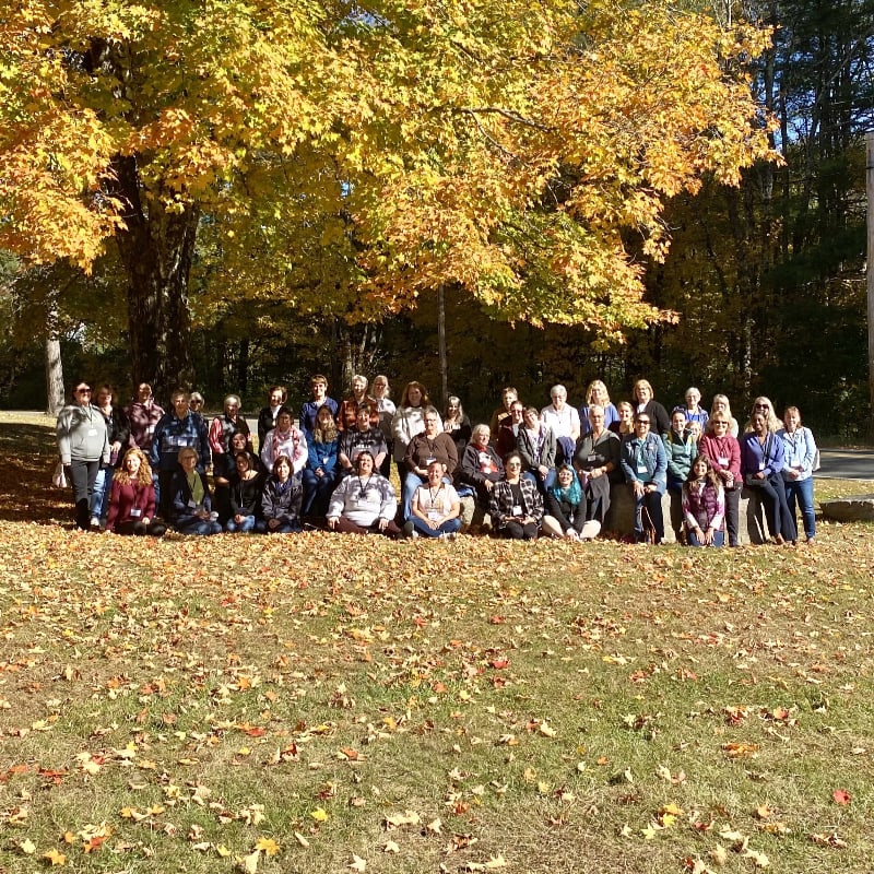 A large group of people pose for a photo in a park with fallen autumn leaves in the foreground and a large tree with golden foliage in the background.