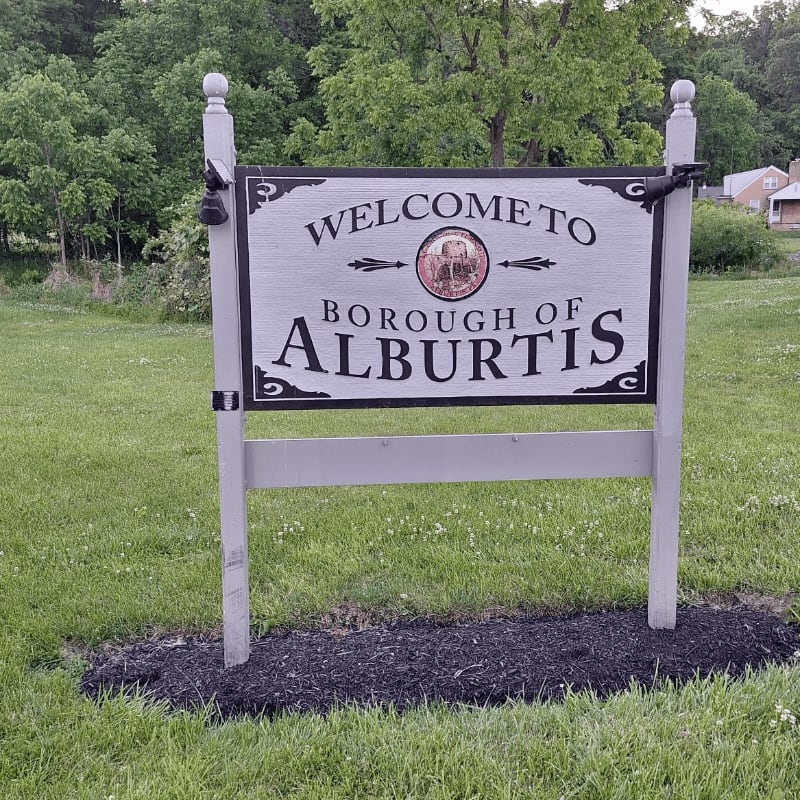 A "Welcome to Borough of Alburtis" sign stands in a grassy area with trees and a house in the background.