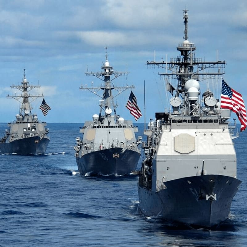 Three US Navy destroyers sail in formation on a calm blue sea under a cloudy sky.