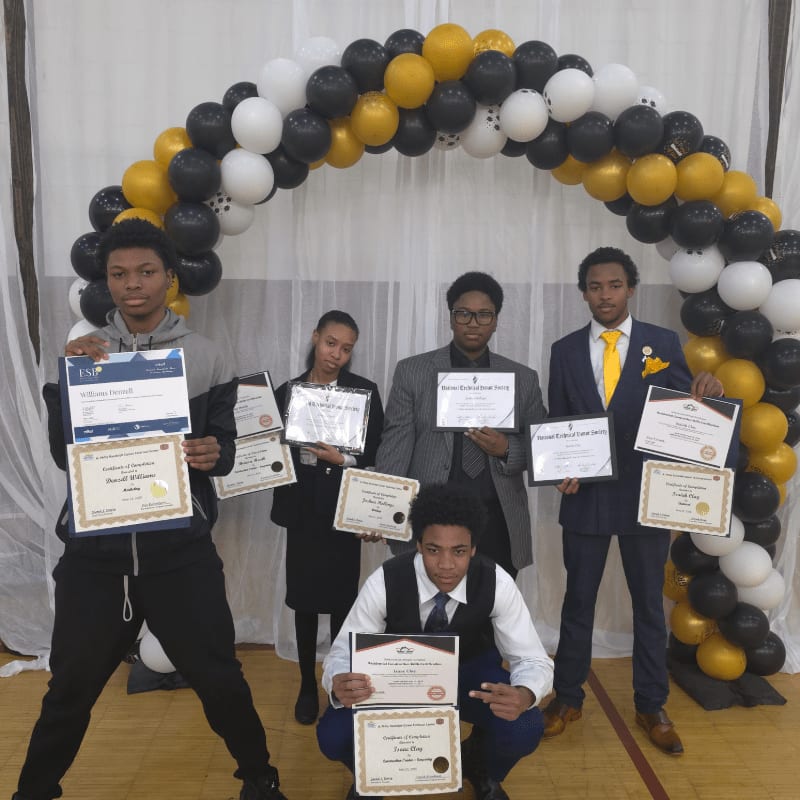 Five young people proudly display their certificates in front of a balloon arch and draped fabric.