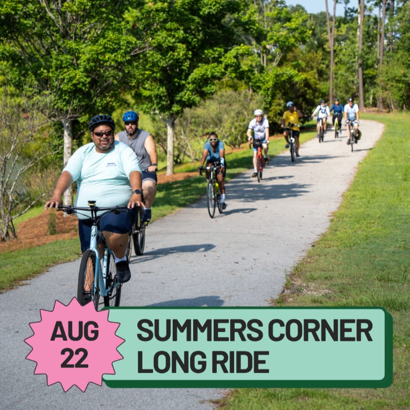 A group of cyclists ride along a paved path in a park, with lush green trees and foliage in the background.