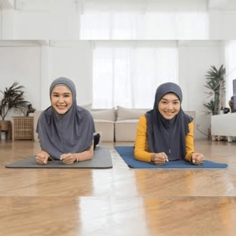 Two smiling women in hijabs are doing planks on yoga mats in the foreground, with a living room and plants visible in the background.