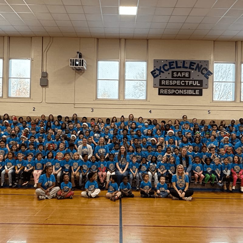 A large group of children and adults in blue shirts are gathered in a gymnasium with a large "EXCELLENCE" sign in the background.