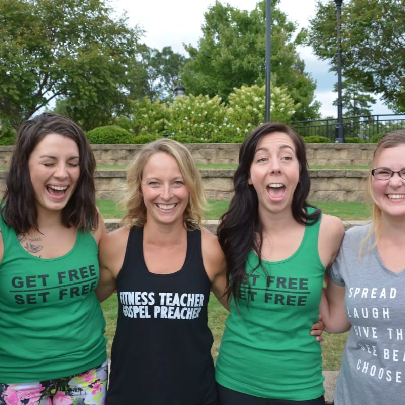 Four women in casual attire smile and laugh in front of a blurred background of trees and greenery.