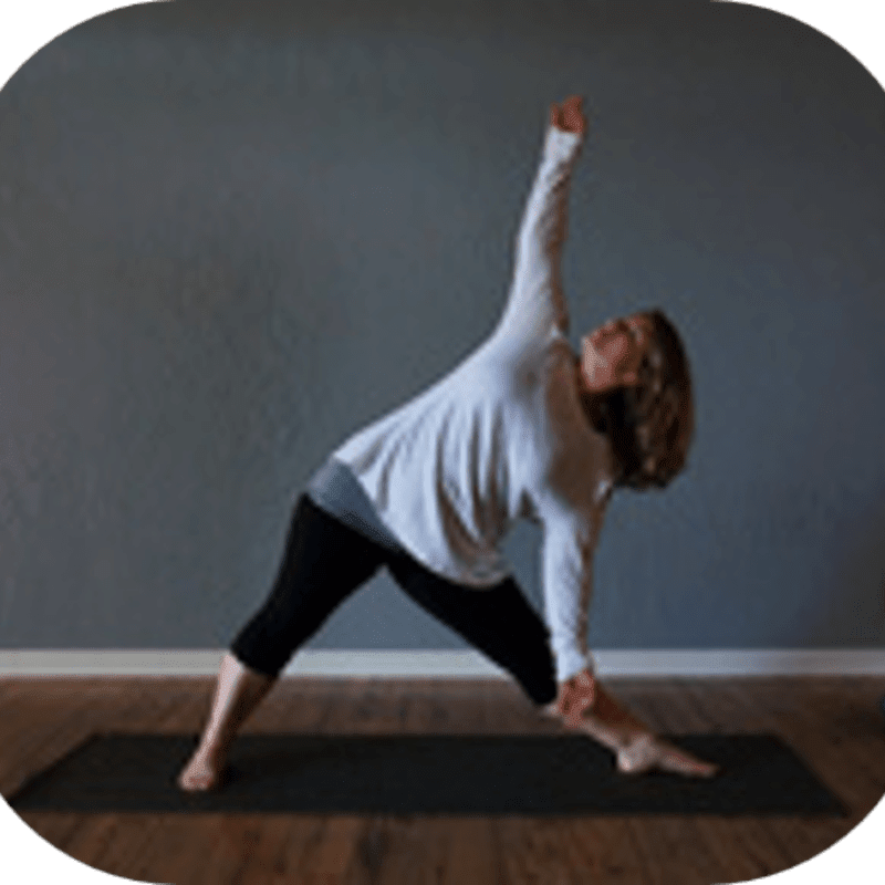 A woman in a white shirt and black leggings performs a triangle pose on a yoga mat in front of a gray wall.