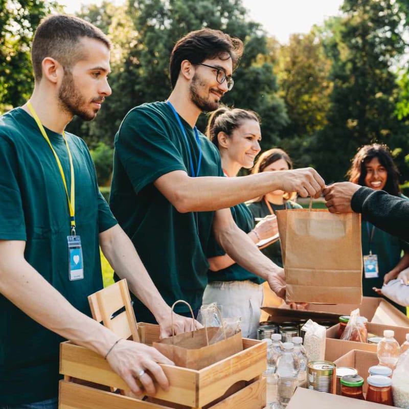 Volunteers in green shirts hand out food items from boxes in the foreground, while a blurred background shows trees and sunlight.