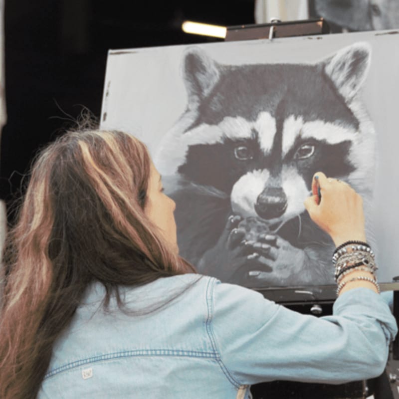 A woman with long, wavy hair and a denim jacket sketches a detailed portrait of a raccoon on an easel, with a dark, blurred background.