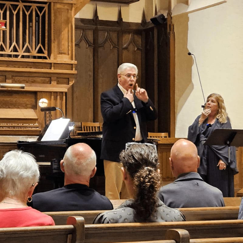 In the foreground, people sit in wooden pews, while in the background, a man and woman sing in front of a piano and ornate wooden paneling.