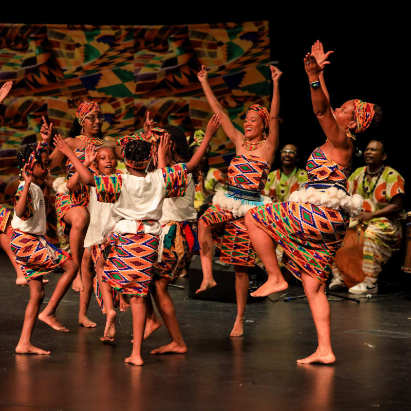 A group of dancers in colorful African attire perform on a stage with a patterned backdrop, while musicians play drums in the background.