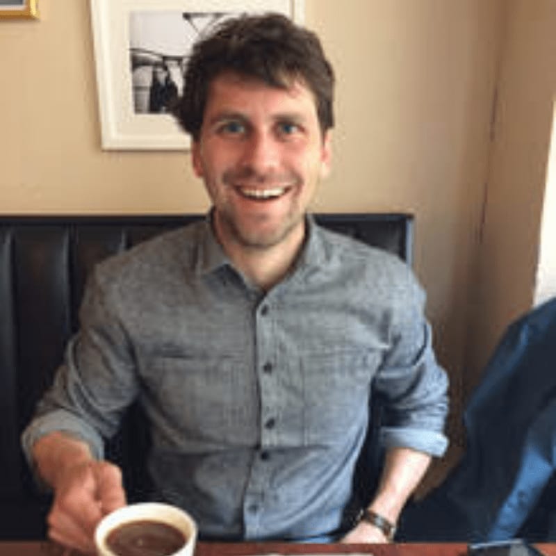 A smiling man in a gray shirt reaches for a cup of coffee in the foreground, with a framed picture on the wall behind him in the background.