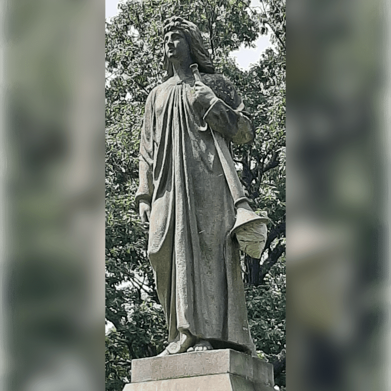 A stone statue of a robed figure holding a palette and a globe stands against a backdrop of lush green trees.