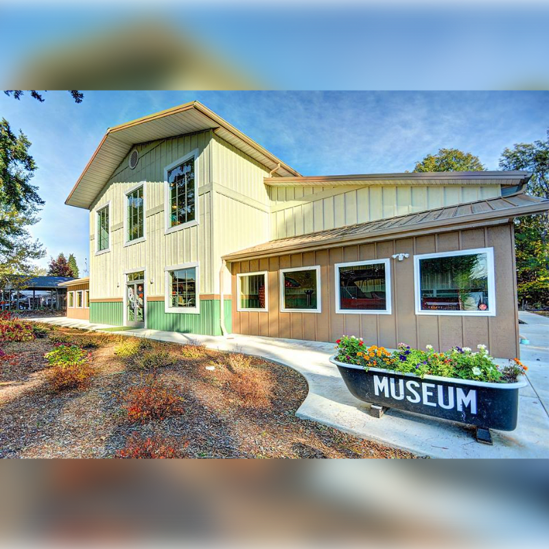 A light green and brown building with a bathtub planter labeled "MUSEUM" in the foreground sits under a bright blue sky with scattered clouds, surrounded by trees and foliage.