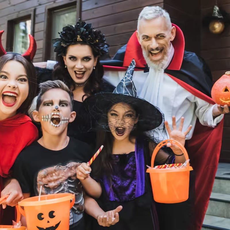 A family in Halloween costumes, including a devil, witch, and vampire, poses with candy buckets in front of a wooden house.