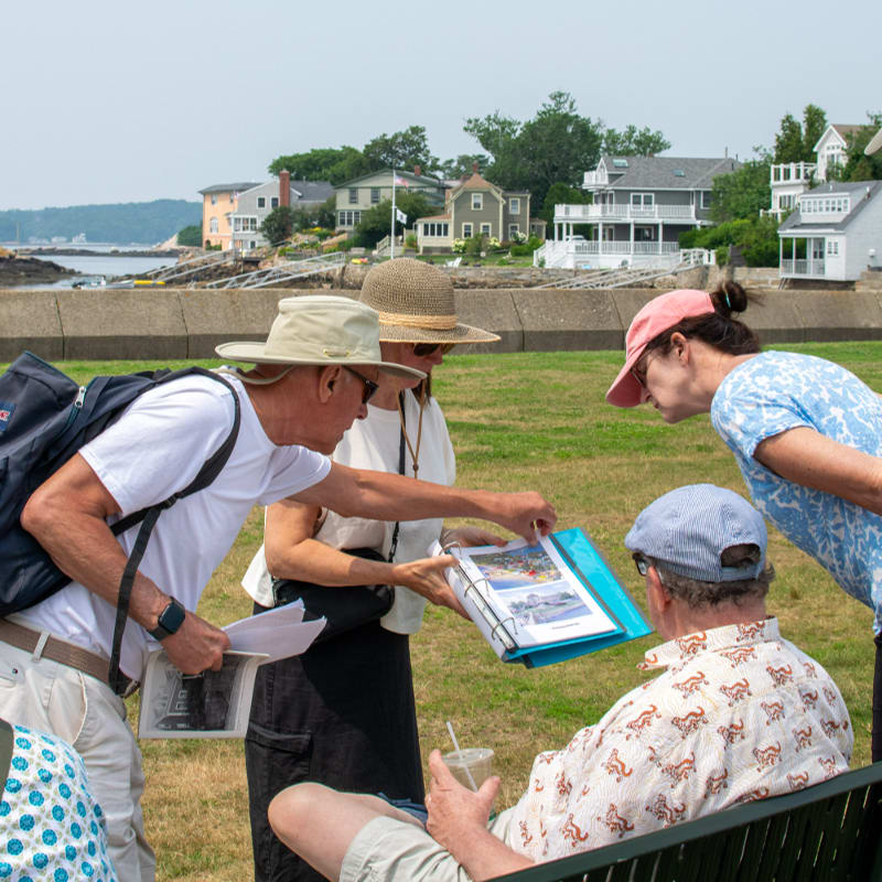 In the foreground, a group of people examine a binder, while in the background, houses line a waterfront under a clear sky.