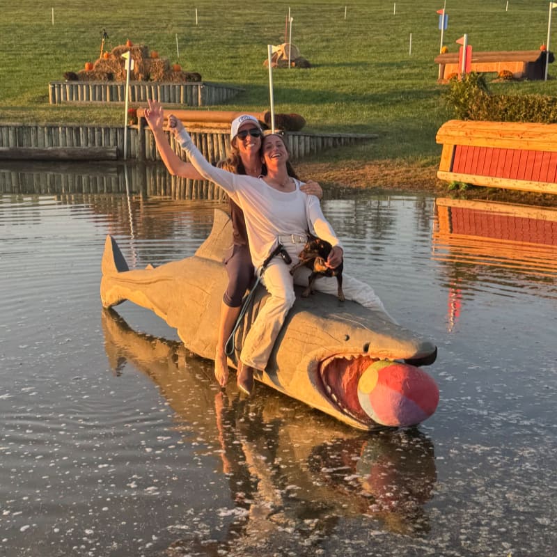 Two women and a dog ride a wooden shark with a ball in its mouth across a calm body of water, with equestrian jumps and a grassy field in the background.