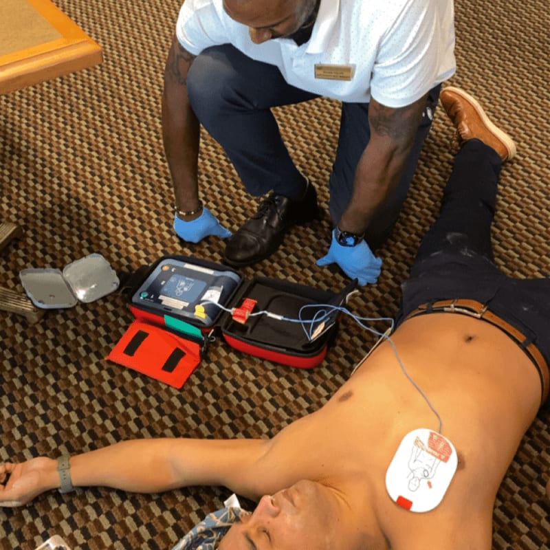 A man in blue gloves kneels beside another man lying on a carpeted floor, operating an automated external defibrillator.