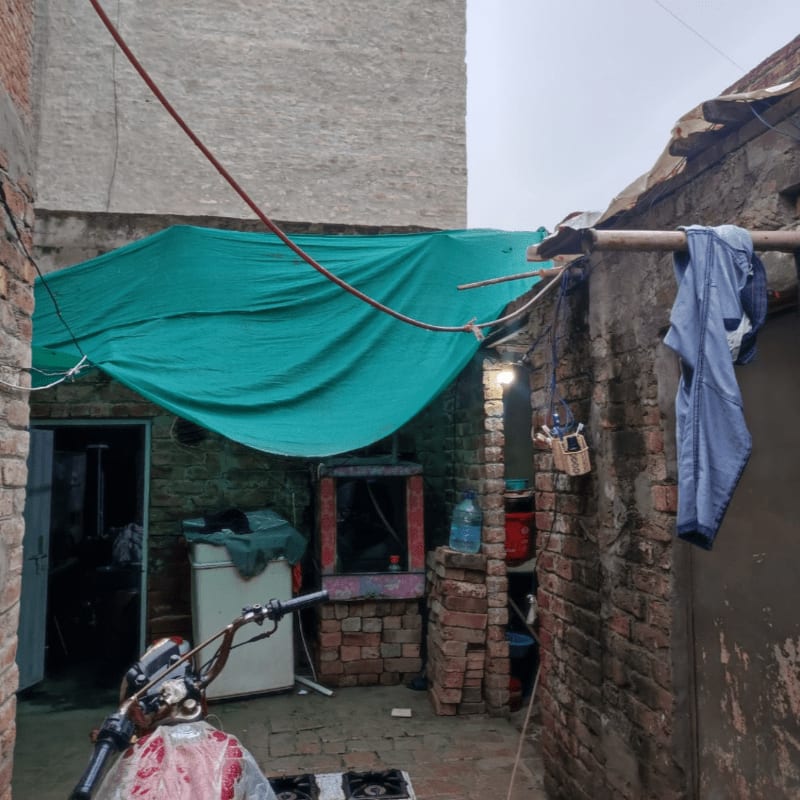 A motorcycle is parked in the foreground of a cluttered courtyard with a green tarp overhead and brick buildings in the background.