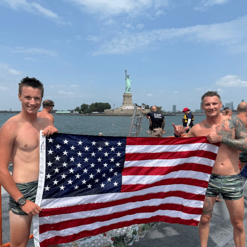 Two men hold an American flag in the foreground with the Statue of Liberty visible in the background across the water.