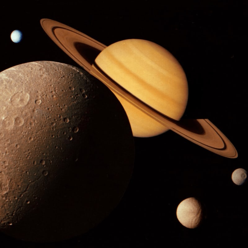 A large, cratered moon dominates the foreground, with Saturn and its rings and several smaller moons visible in the dark, star-filled background.