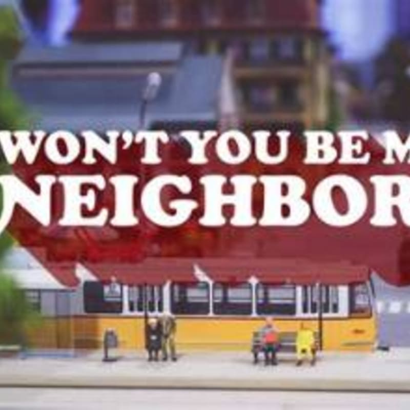 A miniature yellow tram with people on its platform is in the foreground, with buildings and text reading "WON'T YOU BE MY NEIGHBOR" in the background.