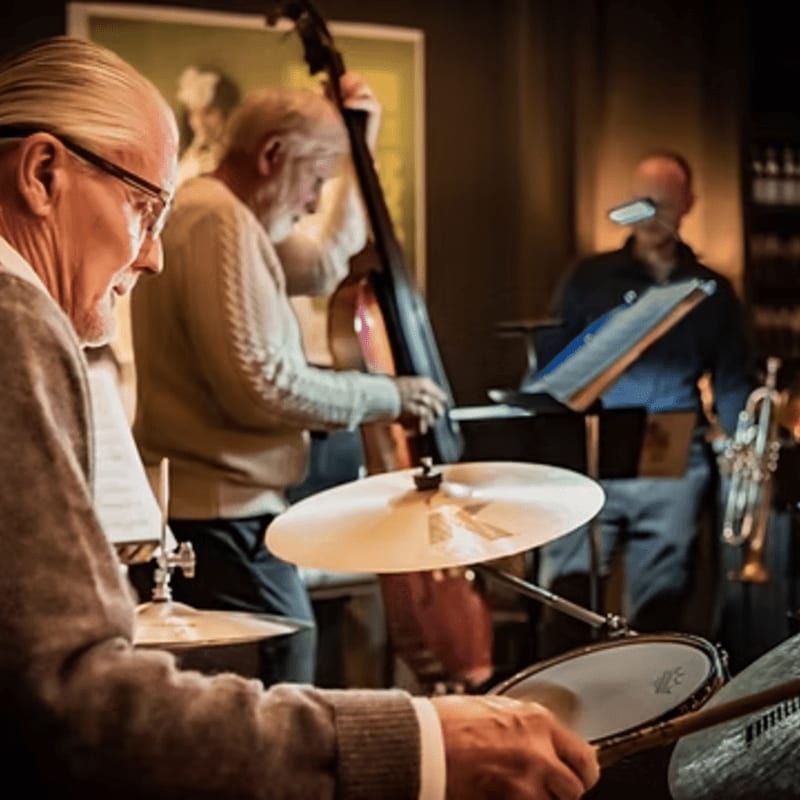 An elderly drummer in the foreground plays his kit while a bassist and another musician perform in the dimly lit background.