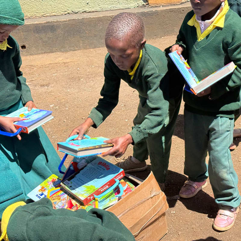 Children in green uniforms sort through a box of books in the foreground, with a dusty, outdoor setting in the background.