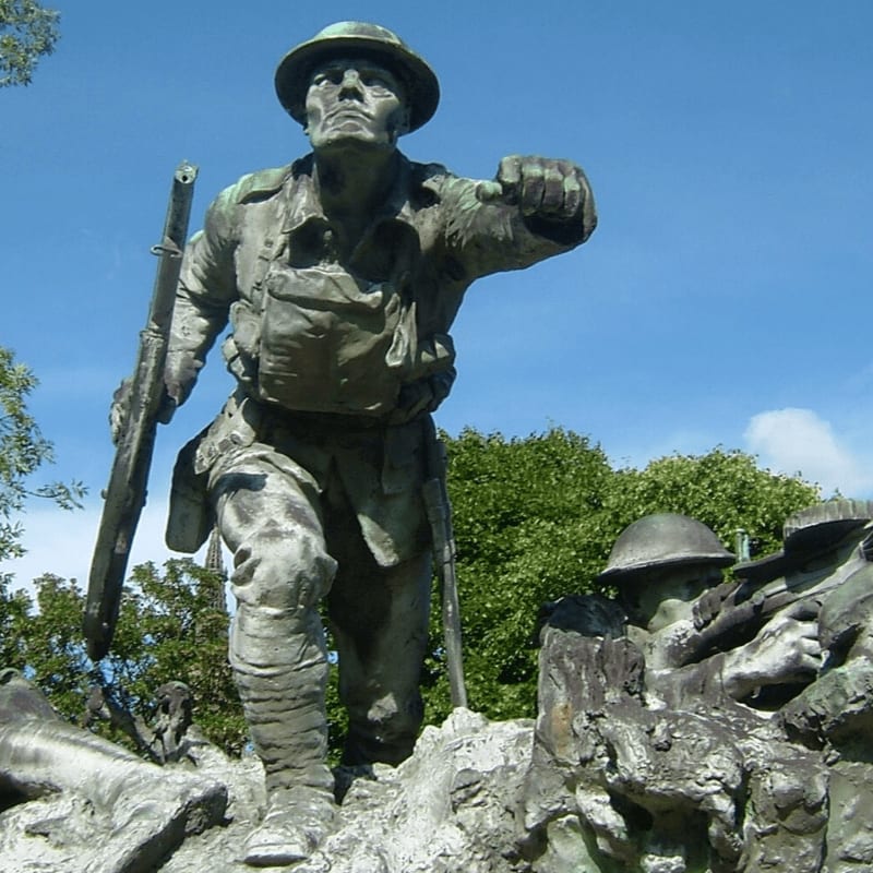A bronze statue of a soldier charging forward with a rifle stands in the foreground against a backdrop of lush green trees and a clear blue sky.