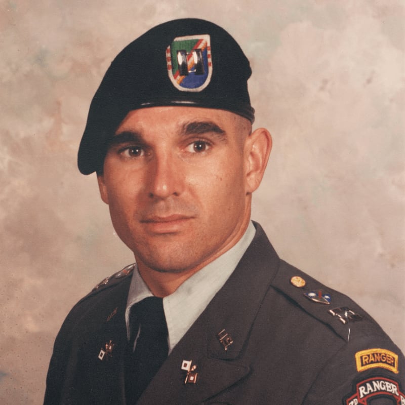 A man in a military uniform with a black beret stands against a mottled background.