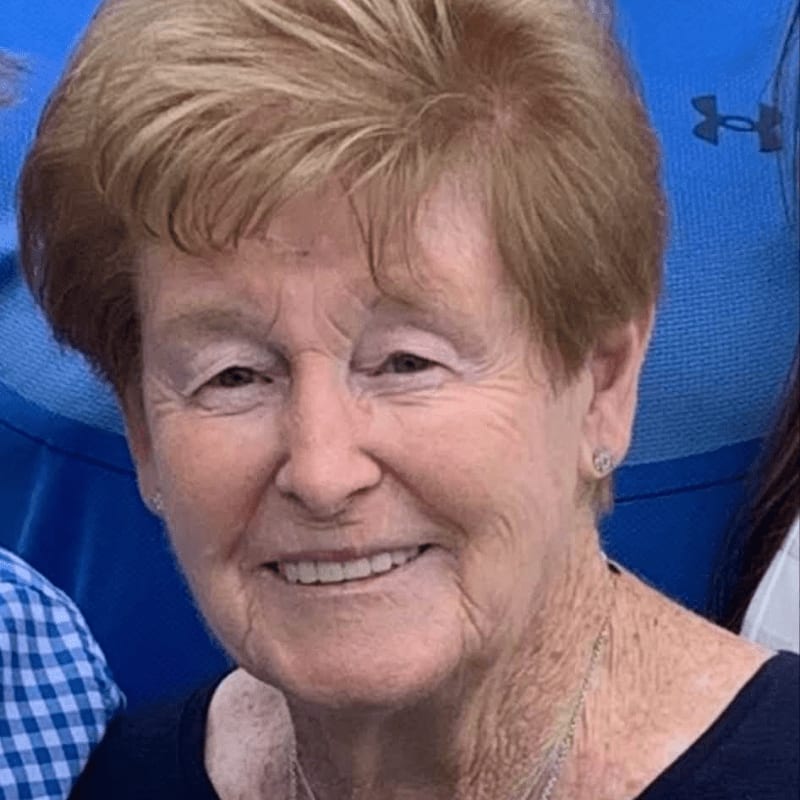 A smiling elderly woman with short blonde hair is in the foreground, with a blurred blue background behind her.