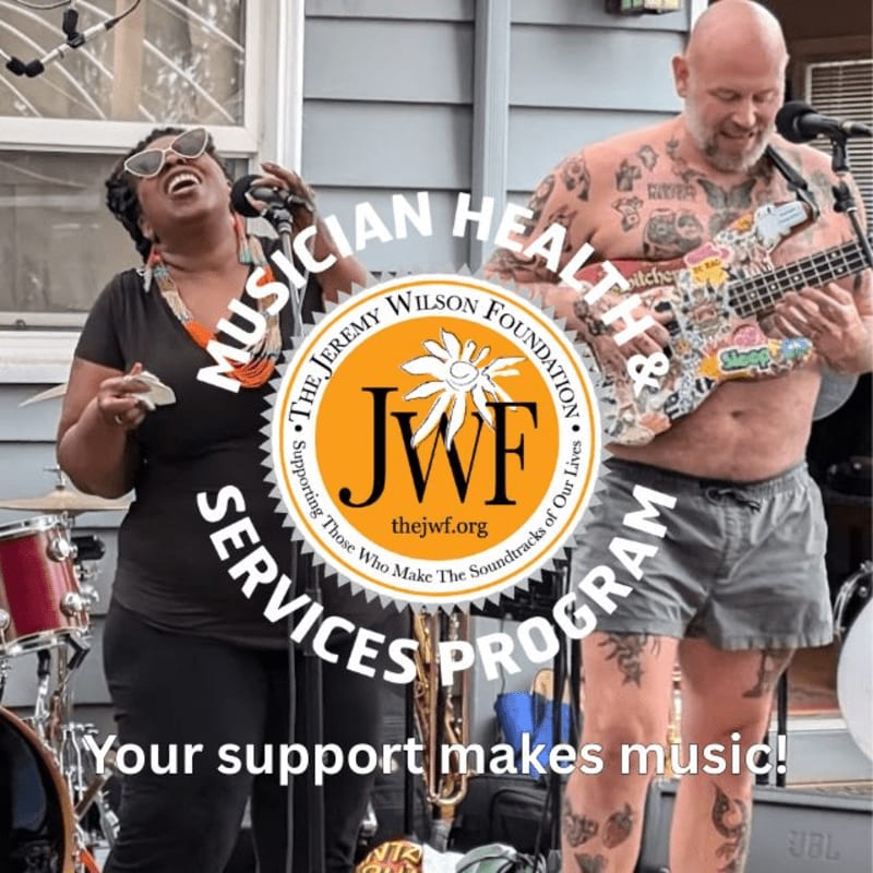 A woman sings into a microphone in the foreground, while a tattooed man plays a guitar in front of a building in the background.