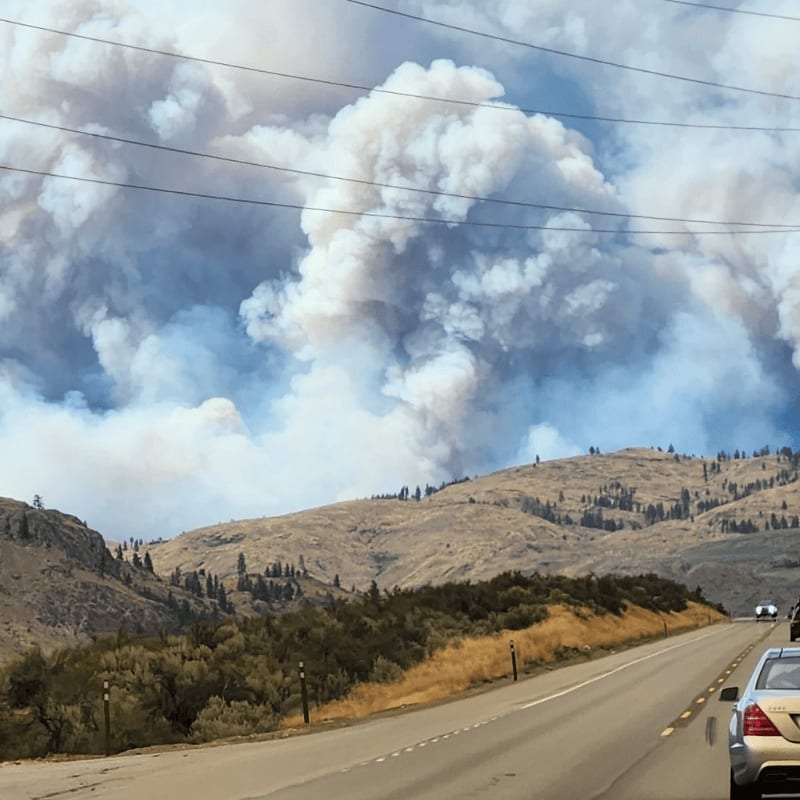 A road curves through a dry, hilly landscape under a sky choked with thick, white smoke from a distant wildfire.