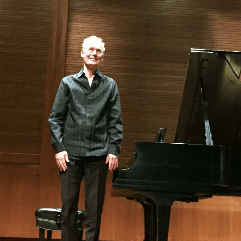 A man in a dark shirt and pants stands next to a grand piano on a stage with a wooden backdrop.
