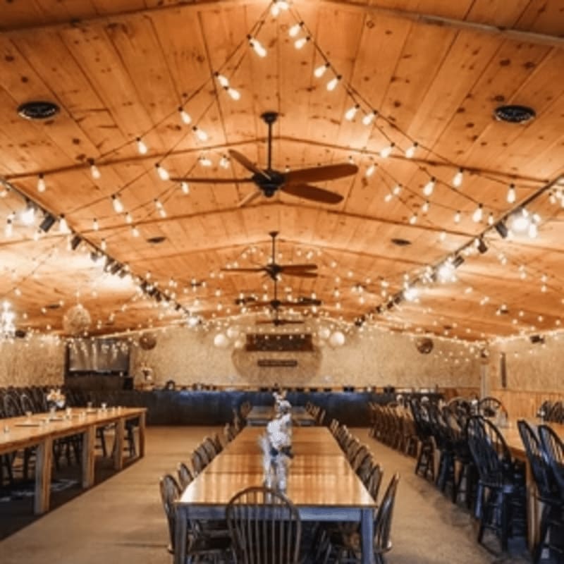 Long wooden tables are set with chairs in the foreground, while the background reveals a warmly lit hall with a vaulted wooden ceiling adorned with string lights.