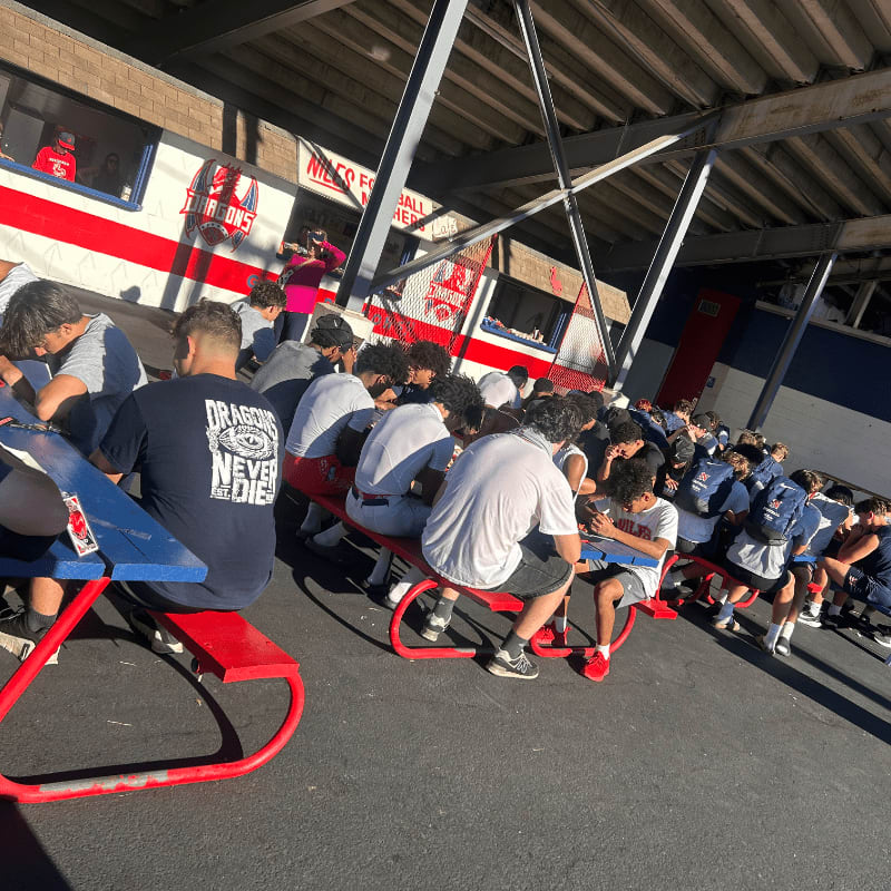 A group of young athletes in the foreground are seated at picnic tables under a covered structure, with a building featuring red and white stripes in the background.