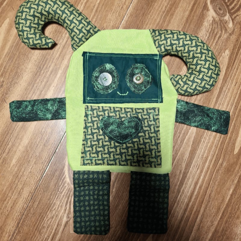 A handmade green stuffed monster with button eyes and a heart on its chest sits on a wooden floor.