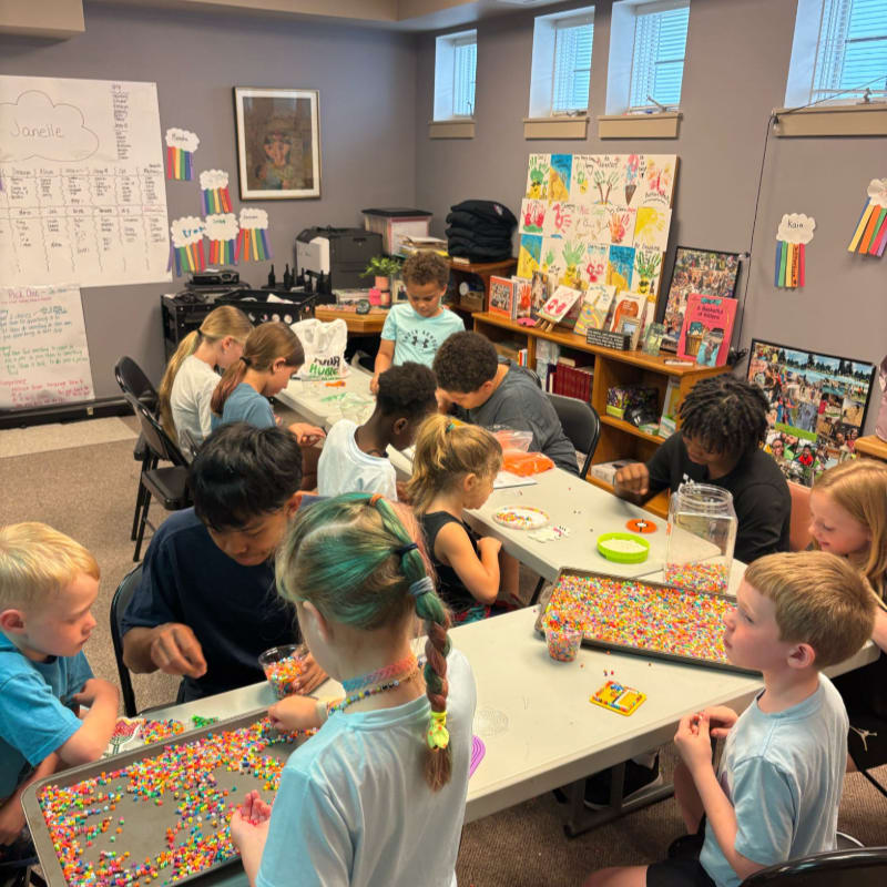 Children are gathered around tables in a classroom, working on a craft project with colorful beads, while a bookshelf filled with books and artwork lines the back wall.