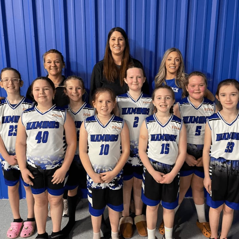 A group of young girls in basketball uniforms stand with their coaches in front of a blue, corrugated metal wall.