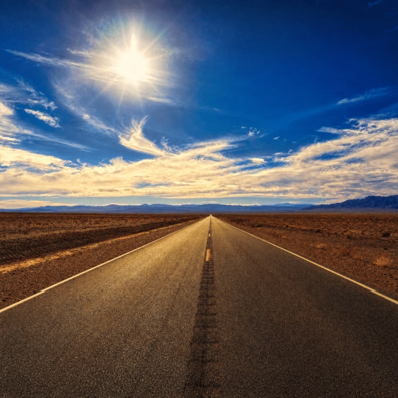 A long, empty asphalt road stretches towards a distant horizon under a bright, sunlit sky with scattered clouds.
