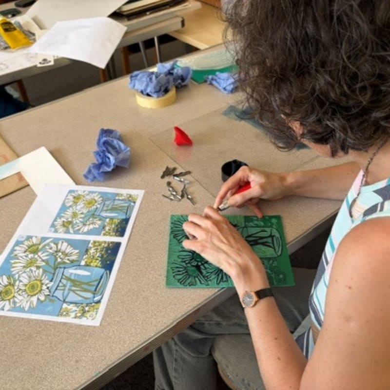 A woman carves a linocut block with sunflowers and vases in the foreground, while a cluttered art studio is visible in the background.