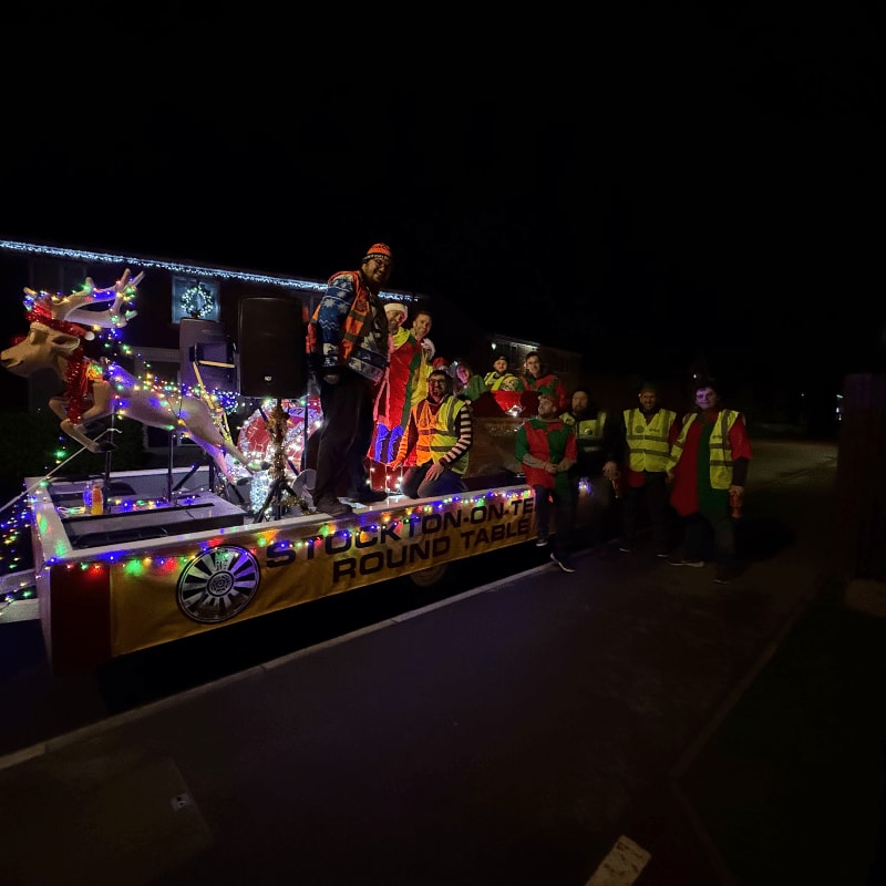 A festive float with people in costumes and a banner reading "STOCKTON-ON-TEES ROUND TABLE" is illuminated by Christmas lights, with a dark, possibly residential, background.