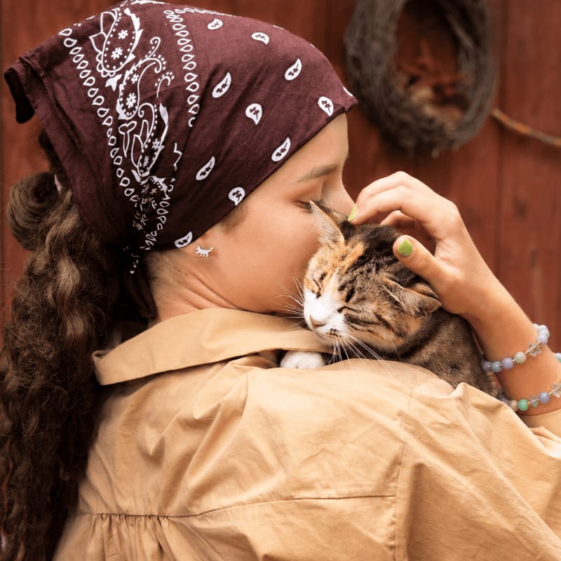 A young woman with a bandana and curly hair gently pets a calico cat nestled on her shoulder, with a rustic wooden wall and a bird's nest in the background.