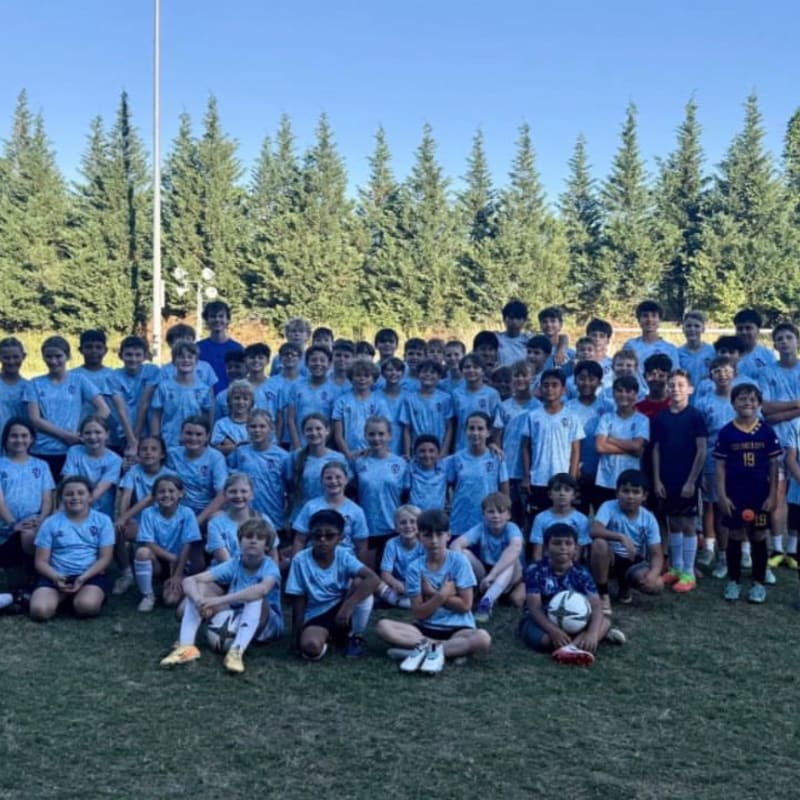 A large group of children in light blue soccer uniforms pose for a photo on a grassy field with a line of evergreen trees in the background under a clear blue sky.