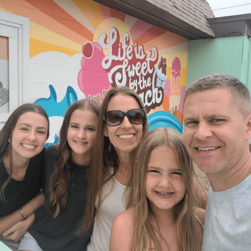A smiling family of five poses for a selfie in front of a colorful mural that reads "Life is Sweet by the Beach."