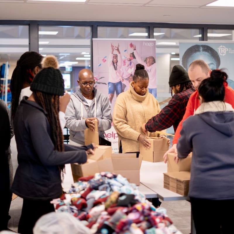 In the foreground, a pile of colorful socks sits next to people packing boxes, while in the background, banners with logos and images are displayed.