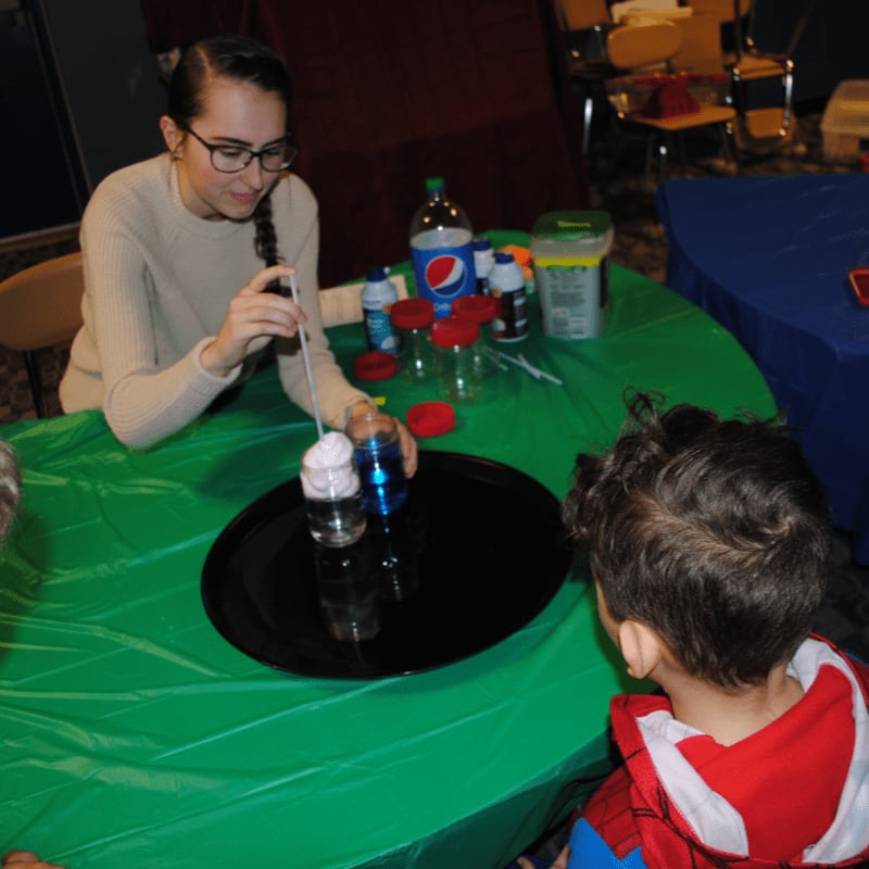 A young woman in glasses demonstrates a science experiment with foam and blue liquid in glasses on a green tablecloth, while a child in a Spider-Man costume watches from the foreground, with chairs and tables visible in the blurred background.