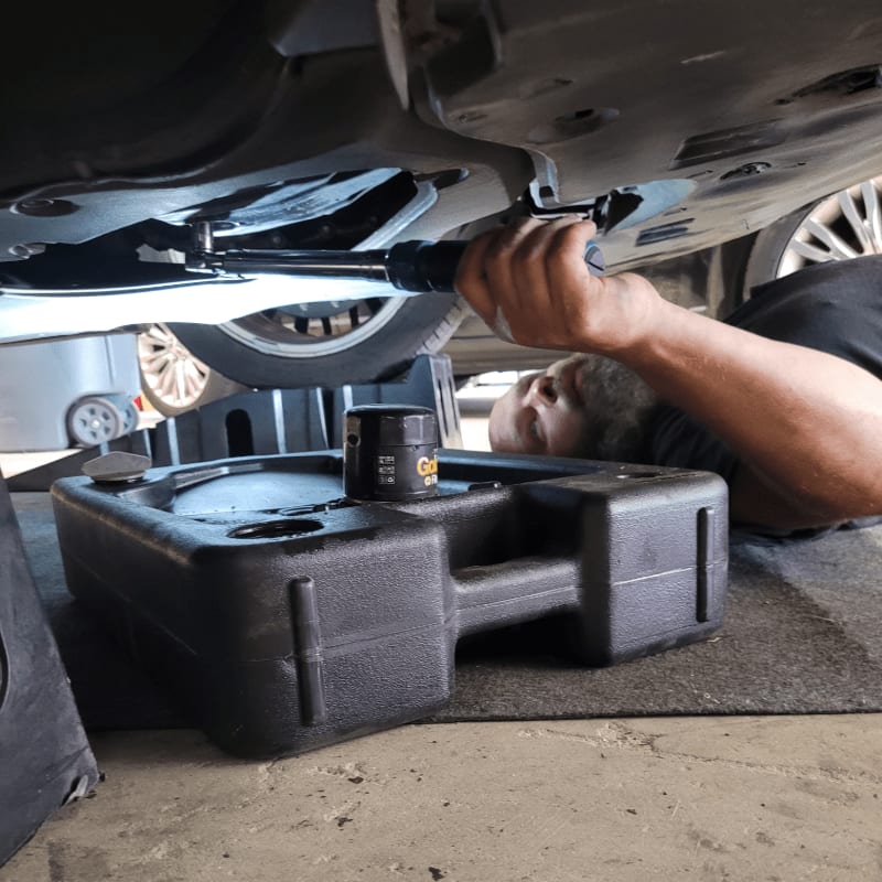 A mechanic uses a wrench to work on a car's undercarriage, with an oil drain pan and filter in the foreground and a car wheel visible in the background.